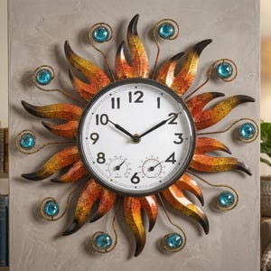 May include: A round wall clock with a white face, black numerals, and two smaller circular gauges at the bottom. The frame features radiating metal sunburst rays in shades of orange, yellow, and brown, accented with eight round blue glass gems.
