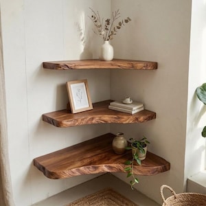 Puede incluir: Tres estantes de esquina escalonados hechos de madera natural con bordes vivos. El estante superior sostiene un jarrón blanco con ramas secas. El estante central presenta una impresión botánica enmarcada y libros. El estante inferior muestra un pequeño jarrón de cerámica y una planta verde colgante.