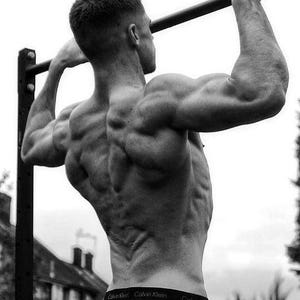 May include: A black and white photograph of a person performing a pull-up on a horizontal metal bar. The person is seen from behind, displaying defined back and arm muscles, wearing light-coloured sweatpants with a black Calvin Klein waistband.
