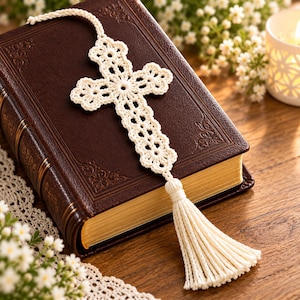 May include: A cream-colored crochet bookmark shaped like a cross with a matching tassel, resting on a dark brown leather-bound book with gold-edged pages. The cross features an intricate openwork floral pattern made of fine thread.