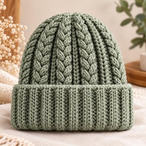 May include: A sage green knit beanie featuring a thick, folded ribbed brim and a vertical cable knit pattern that extends from the crown to the base of the hat. The yarn has a textured, chunky appearance with a consistent matte finish.