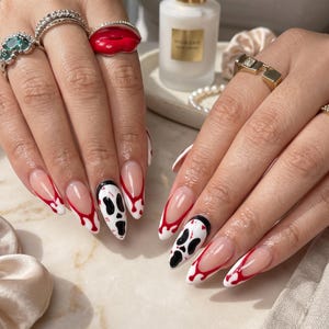 Könnte beinhalten: Mandelformige Nägel im French-Manicure-Stil mit roten blutropfenartigen Spitzen und zwei Akzentnägeln mit einem schwarz-weißen Geistermasken-Design. Die Finger sind mit verschiedenen gold- und silberfarbenen Ringen geschmückt.