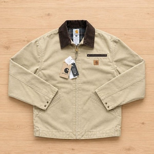 May include: A tan canvas jacket featuring a dark brown corduroy collar, a full-length front zip, and a small zipped chest pocket. A square Carhartt brand patch is visible on the chest, and two paper tags are attached to the zip pull.