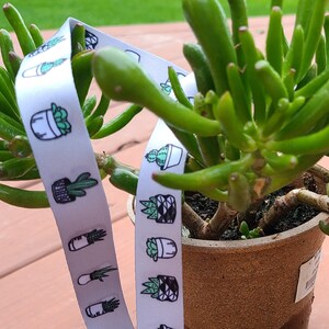 May include: A white lanyard with a repeating pattern of potted cacti and succulents in black and green. The lanyard has a silver-colored metal clasp. The background features a green succulent plant in a brown pot.