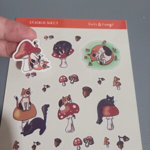 May include: A sticker sheet featuring a selection of charming cat and mushroom illustrations. The sheet includes a black cat, a calico cat, and a mushroom with red and white spots. The text "Cats & Fungi" is printed at the top of the sheet.