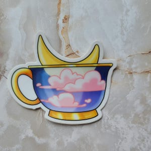 Puede incluir: Una taza de té azul y dorada con una luna creciente y nubes rosadas en su interior. La taza de té tiene un asa dorada y un borde dorado.