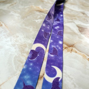 May include: A lanyard with a repeating pattern of manta rays and crescent moons against a starry night sky background. The lanyard is primarily purple and blue, with white accents. The design is printed on a fabric material.