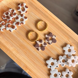May include: A collection of flower-shaped charms and two gold-toned hoop earrings. The charms feature white surfaces with brown spots or brown surfaces with white spots. The earrings are circular with a smooth metallic finish and a hinged clasp.