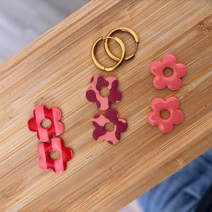May include: A set of jewellery components including two gold-toned metal hoop earrings and six flower-shaped charms. The charms feature three distinct patterns: two with red vertical stripes, two with a dark pink cow print, and two solid coral pink.
