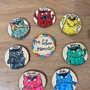 May include: A set of eight circular wooden slices featuring hand-painted monsters representing emotions. The centre piece reads The Colour Monster. Surrounding pieces depict monsters labelled Angry, Happy, Confused, Fear, Calm, Loved, and Sad.