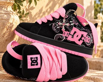 May include: A pair of black and pink DC sneakers featuring thick pink laces, a pink DC logo on the side and tongue, and a silver cross charm with rhinestones attached to the laces. The black fabric upper is decorated with a white swirling pattern.