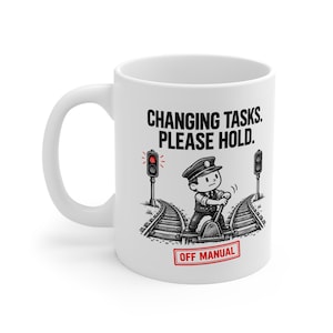 May include: A white ceramic mug featuring a black and white illustration of a train conductor operating a track switch. The text reads "CHANGING TASKS. PLEASE HOLD." at the top and "OFF MANUAL" in a red rectangular outline at the bottom.