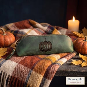 May include: A dark green leather pencil case featuring a centered embossed pumpkin graphic with the letter M inside. The case has a gold-toned zipper closure with a matching leather pull tab. The text Design H2 Pumpkin Initial is visible.