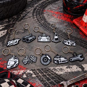 Peut inclure: Un ensemble de dix porte-clés en caoutchouc noir et blanc sur le thème automobile, incluant le mot DRIFT, des pots d'échappement, des voitures de sport, un cône de signalisation, un casque, des drapeaux à damier, un disque de frein et une clé.