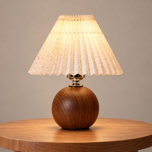 May include: A table lamp featuring a spherical dark brown wood base with a visible grain pattern and a metallic silver neck. The lamp is topped with a conical, pleated, light beige fabric lampshade that emits a warm, soft glow when illuminated.