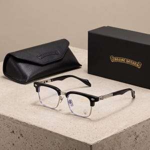 May include: Black and silver-toned spectacles featuring a browline frame design with metallic temple detailing and a fleur-de-lis emblem on the lens. Includes a black textured leather protective case and a black box with the Chrome Hearts logo.