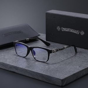 Puede incluir: Gafas rectangulares negras con detalles metálicos en tono plateado en las patillas y un pequeño emblema de cruz en el cristal. La montura viene acompañada de un estuche de cuero negro y una caja negra, ambos con la marca Chrome Hearts.