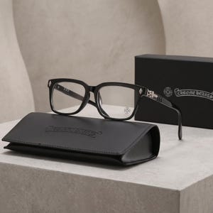 Può includere: Occhiali con montatura nera e lenti trasparenti, caratterizzati da dettagli in metallo color argento sulle aste e un piccolo emblema a croce sulla lente. Gli occhiali sono presentati con una custodia in pelle nera e una scatola nera, entrambe con il logo Chrome Hearts.