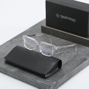 May include: Clear rectangular eyeglasses with silver-toned ornate metal temples featuring intricate engravings. The glasses are accompanied by a black leather case and a black box, both displaying the Chrome Hearts logo in silver lettering.