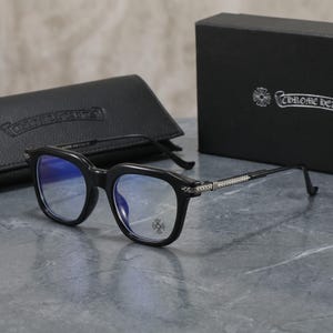 May include: Black-framed spectacles with blue-tinted lenses and silver-toned metallic temple arms featuring intricate detailing. A small cross emblem is visible on the right lens. Includes a black leather-textured case and a black box with text.