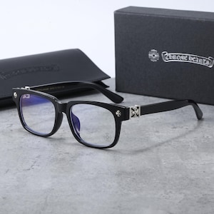 May include: Black rectangular spectacles featuring silver-toned cross emblems on the temples and hinges. The lenses have a blue light tint, and a small cross logo is etched on the right lens. The glasses are accompanied by a black case and box.