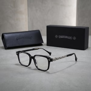 May include: Black eyeglasses with clear lenses and rectangular frames. The temples feature intricate silver-toned metal detailing with ornate patterns. A black leather-like case and a black rectangular box, both marked with Chrome Hearts, are included.