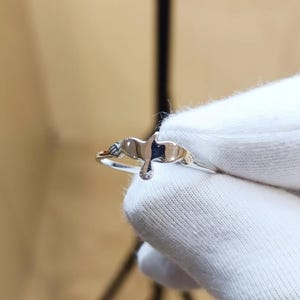 May include: A polished silver-toned ring featuring a central bird-shaped motif with outstretched wings. The band is thin and metallic, showcasing a smooth, reflective surface that catches the light, creating a clean and simple design.