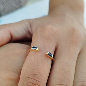 Könnte beinhalten: Ein offener Goldring mit zwei im Baguetteschliff geschliffenen blauen Edelsteinen an den Enden der Ringschiene. Der Ring hat eine polierte Goldoberfläche und die beiden rechteckigen blauen Steine liegen sich gegenüber.