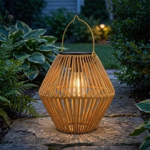 Op de afbeelding: Een lantaarn met een lichtbruine, kooivormige structuur van dunne, verticale staven. De lantaarn heeft een handvat aan de bovenkant en een gloeiende lamp binnenin, wat zorgt voor warme verlichting. Het heeft een donker, rond paneel aan de bovenzijde.