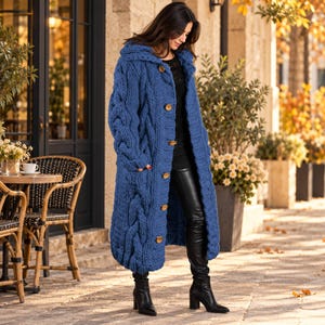 May include: A long, blue, chunky cable-knit cardigan featuring a hood, long sleeves, and a front button closure with five large, round, brown buttons. The garment is worn over a black top and black pants with black high-heeled boots.