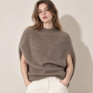 Könnte beinhalten: Ein taupefarbener, flauschiger, ärmelloser Strickpullover mit Stehkragen, kombiniert mit einer weißen Jeanshose. Der Pullover hat eine weiche, gebürstete Textur und eine lockere, entspannte Passform, die über Schultern und Rumpf fällt.