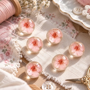 May include: A set of five spherical clear resin buttons, each containing a preserved pink cherry blossom flower. The buttons are arranged on a piece of fabric, with a wooden spool of pink thread and a pair of gold scissors visible nearby.