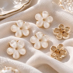 May include: A collection of flower-shaped buttons featuring a mix of translucent white petals with subtle glitter accents and gold-toned metal centers, alongside gold-toned metal flower buttons with intricate textured petal details.