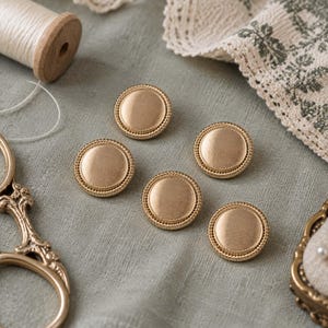 May include: Five circular gold-toned buttons with a brushed metallic finish and a beaded edge. The buttons are arranged in a cluster, each featuring a smooth, slightly concave center and a textured, decorative border around the circumference.