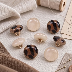 May include: A collection of seven round shank buttons featuring a mix of solid cream-colored designs and brown leopard print patterns. Each button has a smooth, glossy finish and a gold-toned metal loop on the underside for attachment.