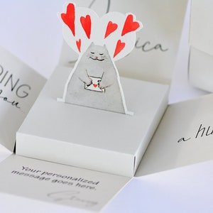 May include: A pop-up card with a gray cat holding a red heart-shaped envelope. The card is open and the inside reads "Sending you a hug." The card is decorated with red hearts.