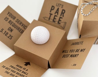 May include: A golf-themed gift box with a white golf ball at the center. The box is brown with black text that reads "Let's Par Tee" and "Will you be my best man?" A practice ball is included.