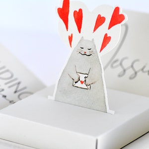 May include: A white pop-up card with a gray cat holding a red envelope with a heart on it. The cat is surrounded by red hearts. The card says "Sending you" on the front and "Jessica" on the back.