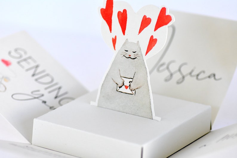 May include: A white pop-up card with a gray cat holding a red envelope with a heart on it. The cat is surrounded by red hearts. The card says "Sending you" on the front and "Jessica" on the back.