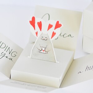May include: A white pop-up card with a cat holding a letter and surrounded by red hearts. The card reads "Sending you a hug."