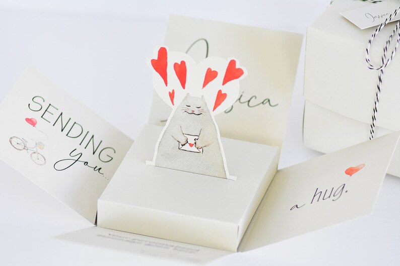 May include: A white pop-up card with a cat holding a letter and surrounded by red hearts. The card reads "Sending you a hug."