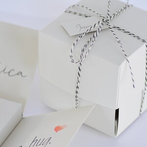 May include: A white gift box tied with black and white twine. The box has a small white tag with the word "Jessica" written on it. The box is sitting on a white surface.