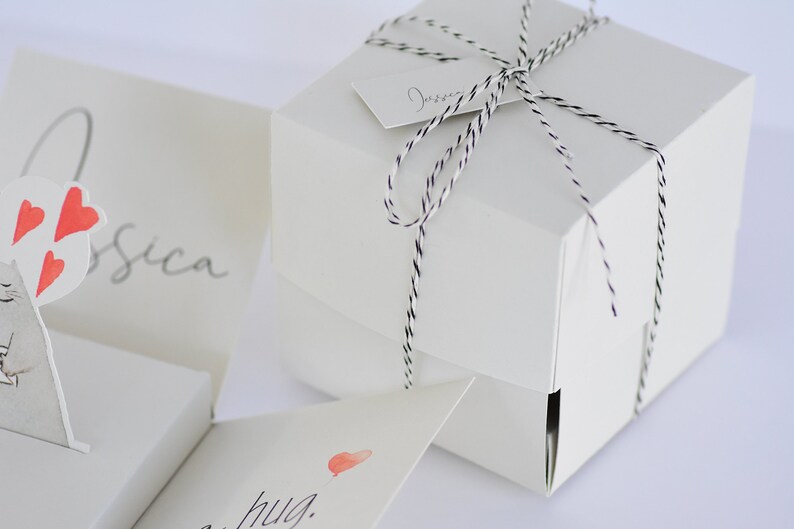May include: A white gift box tied with black and white twine. The box has a small white tag with the word "Jessica" written on it. The box is sitting on a white surface.