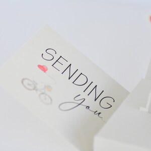 May include: A white card with the text "Sending you..." in black lettering. A small red heart and a bicycle with a red balloon are illustrated on the card.