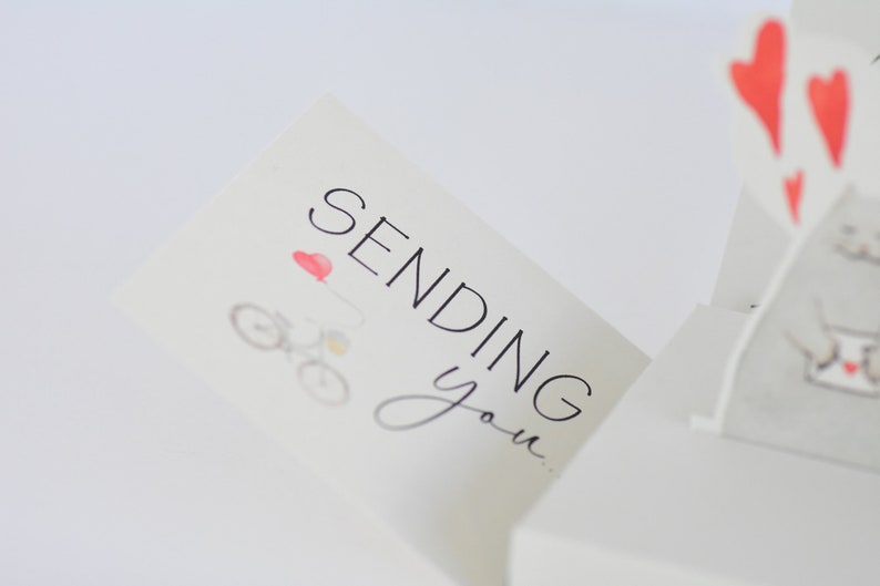 May include: A white card with the text "Sending you..." in black lettering. A small red heart and a bicycle with a red balloon are illustrated on the card.