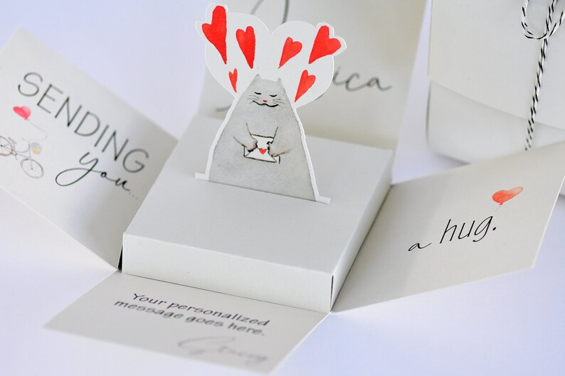 May include: A pop-up card with a gray cat holding a red heart-shaped envelope. The card is open and the inside reads "Sending you a hug." The card is decorated with red hearts.