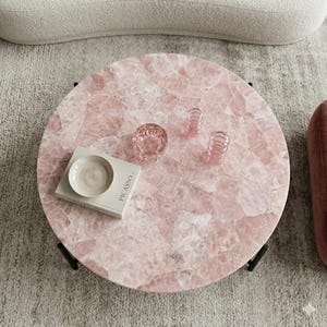 May include: A round coffee table with a pink quartz stone top and black metal legs. On the surface sits a white book titled Picasso, a small white ceramic bowl, and two small pink glass ribbed candle holders arranged in a cluster.