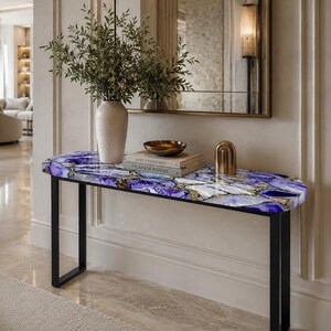 May include: A console table with a rectangular top featuring a purple and white agate stone pattern with gold accents, supported by a black metal frame. On the table are a beige vase with greenery, two stacked books, and a gold arch object.