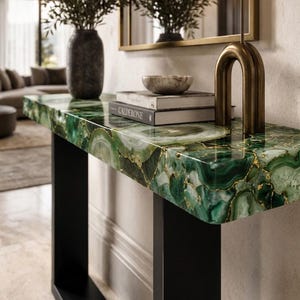 May include: A console table featuring a rectangular top made of polished green agate slices with gold-toned veining, supported by a black metal frame. The table surface holds two stacked books, one labeled Calderone, and a brass sculpture.