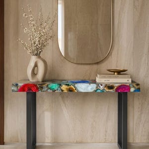 May include: A console table with a multicolored agate stone top featuring vibrant red, blue, green, and yellow hues, supported by two black metal legs. A beige ceramic vase with white flowers and two stacked books sit on the surface.